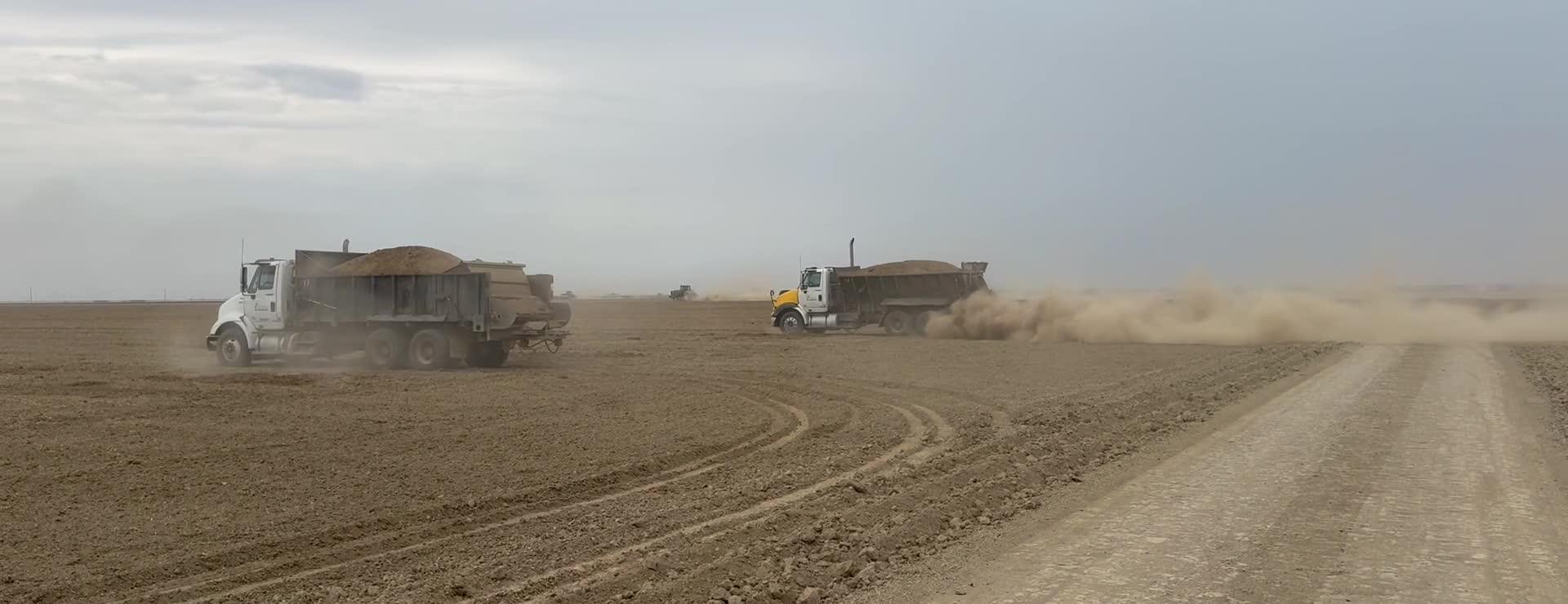 Two B. Mello spreader trucks working open ground, the second pulling away and starting to spread